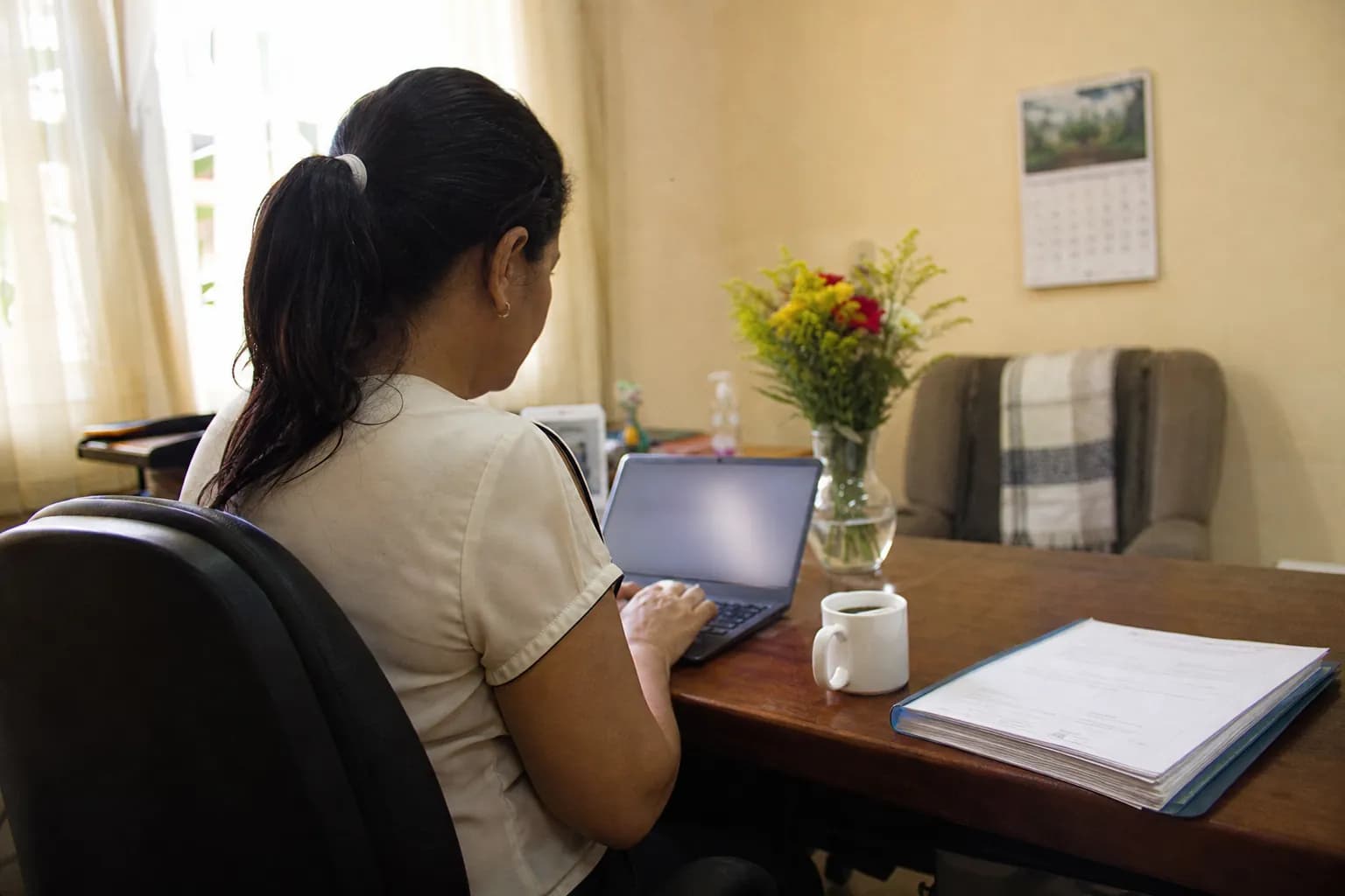 Administradora trabalhando no notebook na sala da administração, com contratos e café sobre a mesa